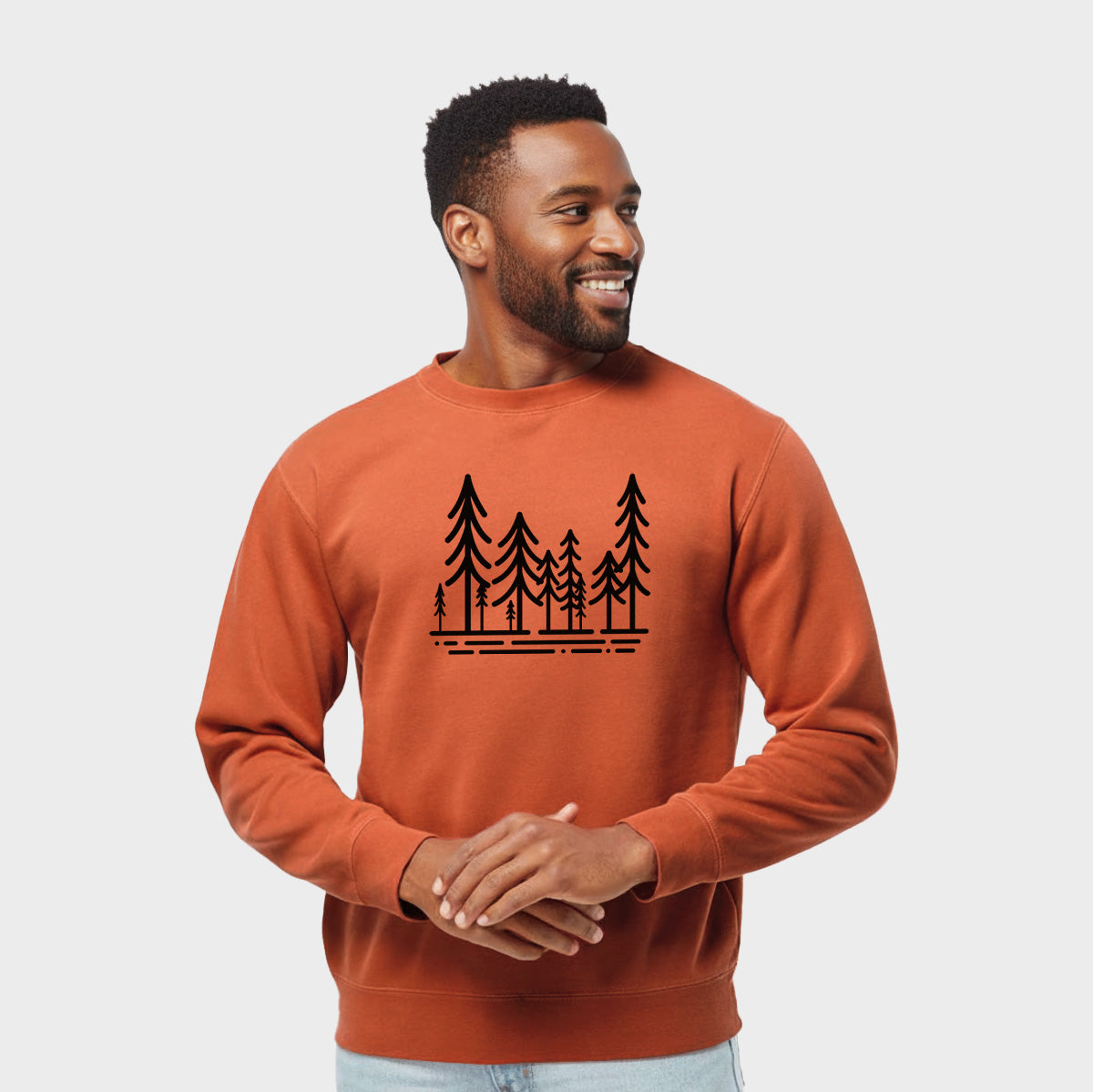 Model wearing an amber orange pigment dyed crewneck sweatshirt printed with bold black line art of ten simple pine trees above horizontal lines.