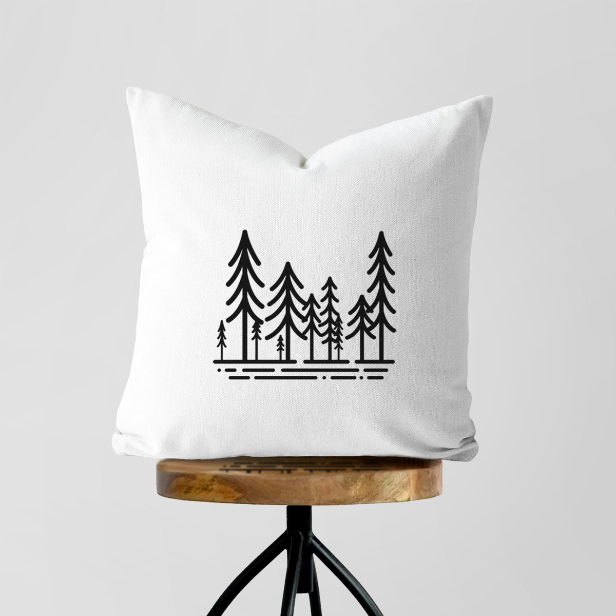 White pillow cover printed with bold black line art of ten simple pine trees above horizontal lines, on a wooden stool.