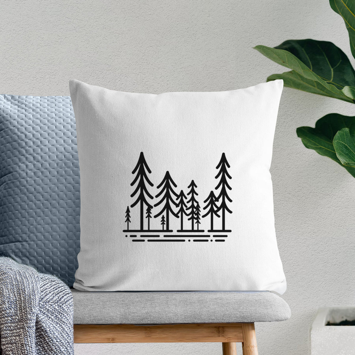 White pillow cover printed with bold black line art of ten simple pine trees above horizontal lines, on a bench beside plants.