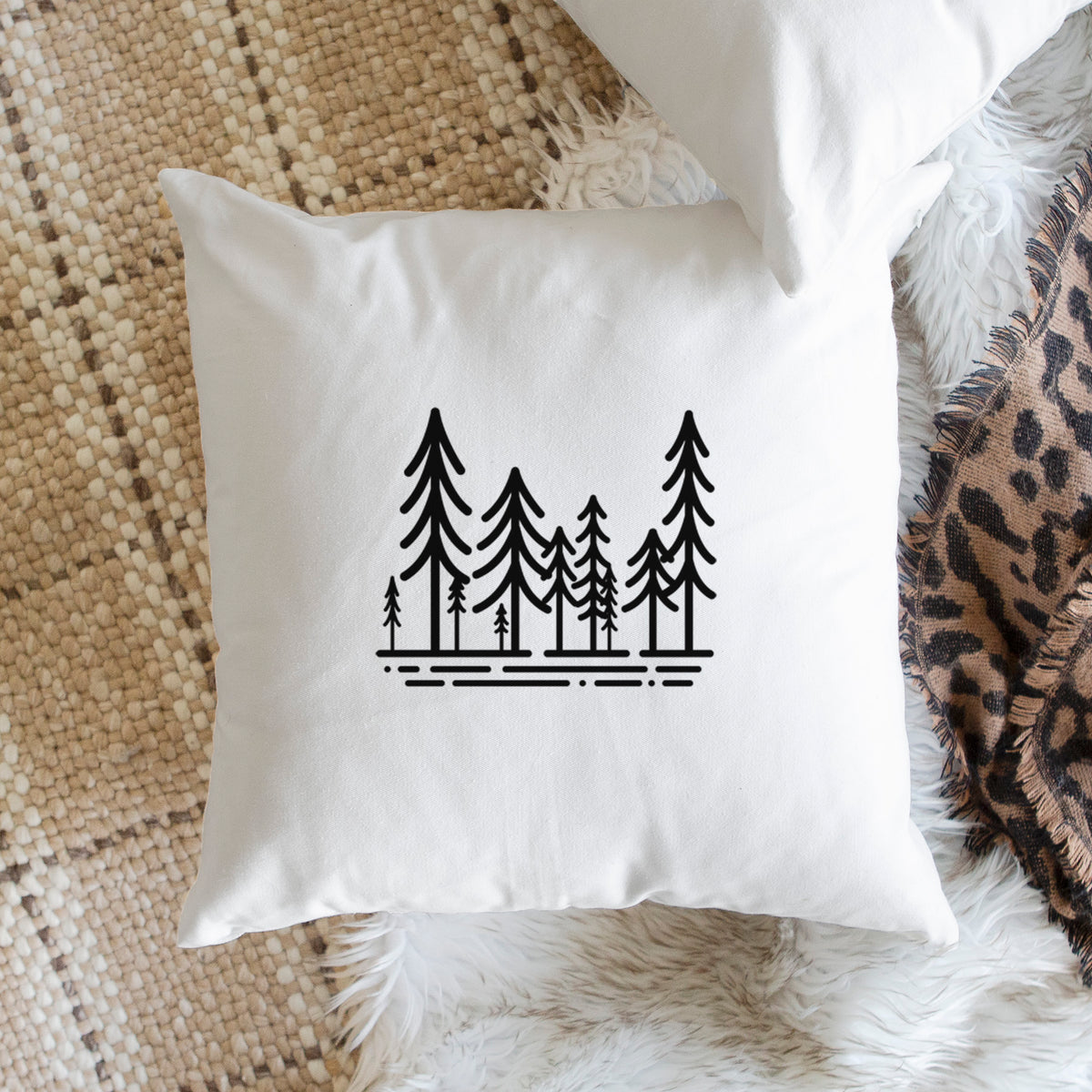 White pillow cover printed with bold black line art of ten simple pine trees above horizontal lines, on a woven chair.