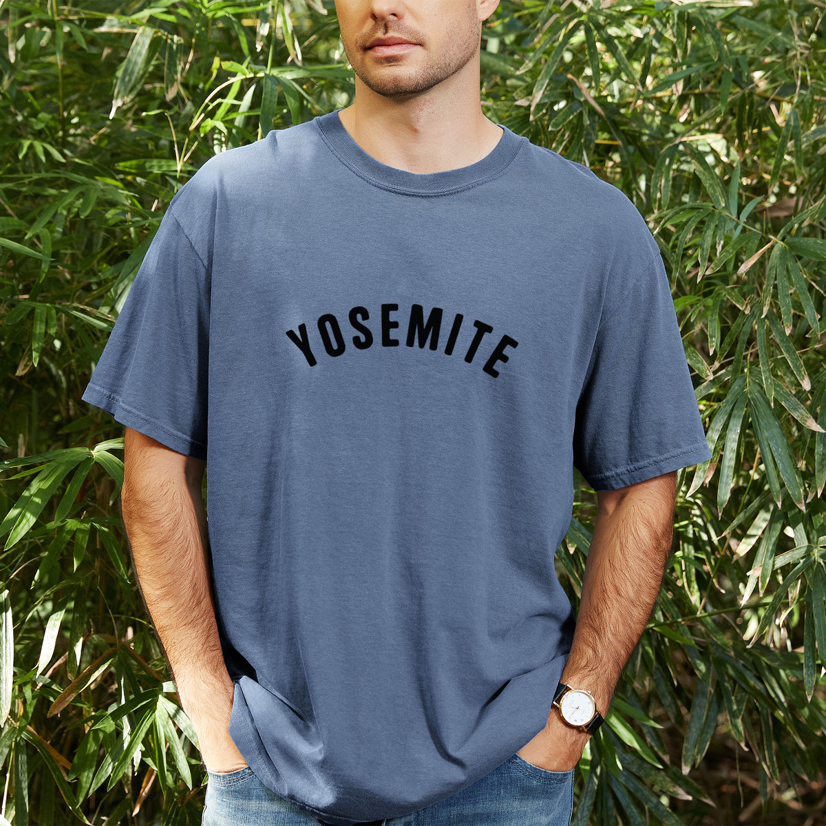 Man wearing a Comfort Colors t-shirt printed with the word Yosemite in bold black letters arched across the chest, outdoors.