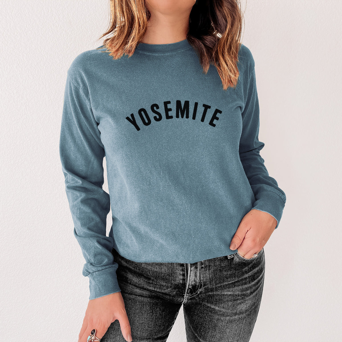 Steel blue Comfort Colors long sleeve shirt printed with the word Yosemite in bold black letters arched across the chest, shown in a lifestyle photo.
