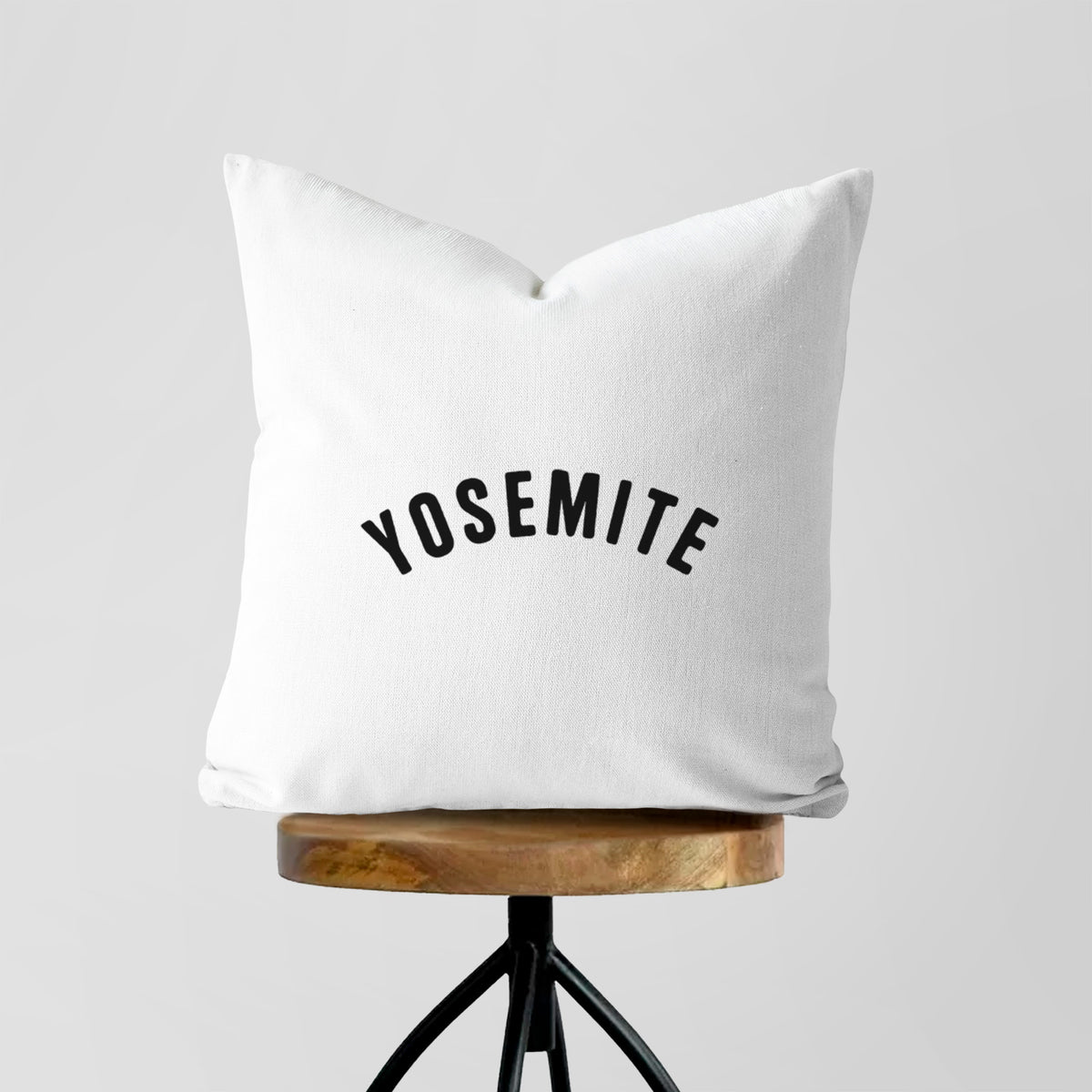 White pillow cover printed with the word Yosemite in bold black letters arched across the chest, on a wooden stool.