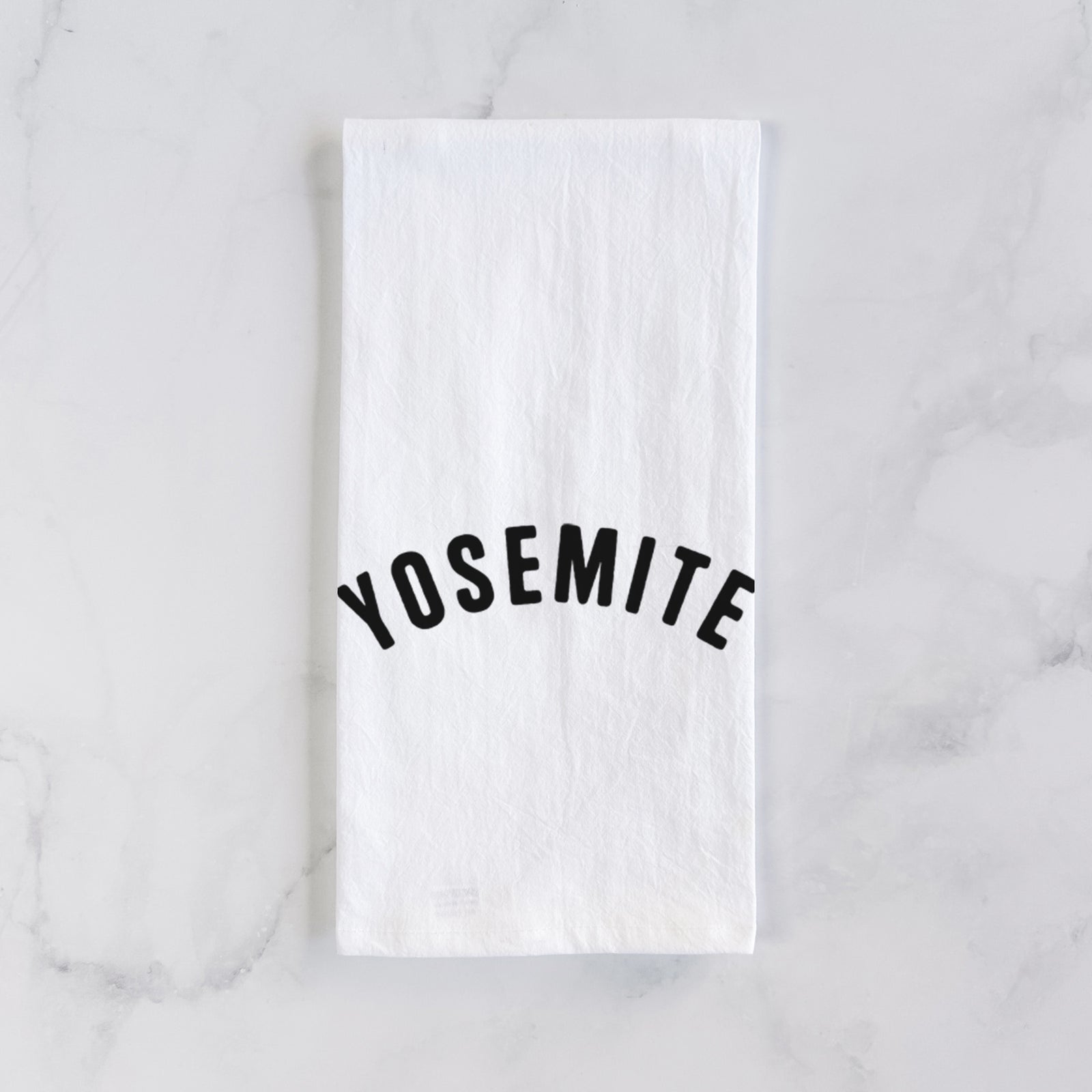 White cotton tea towel printed with the word Yosemite in bold black letters arched across the chest, on a marble counter beside a round wooden cutting board and greenery.