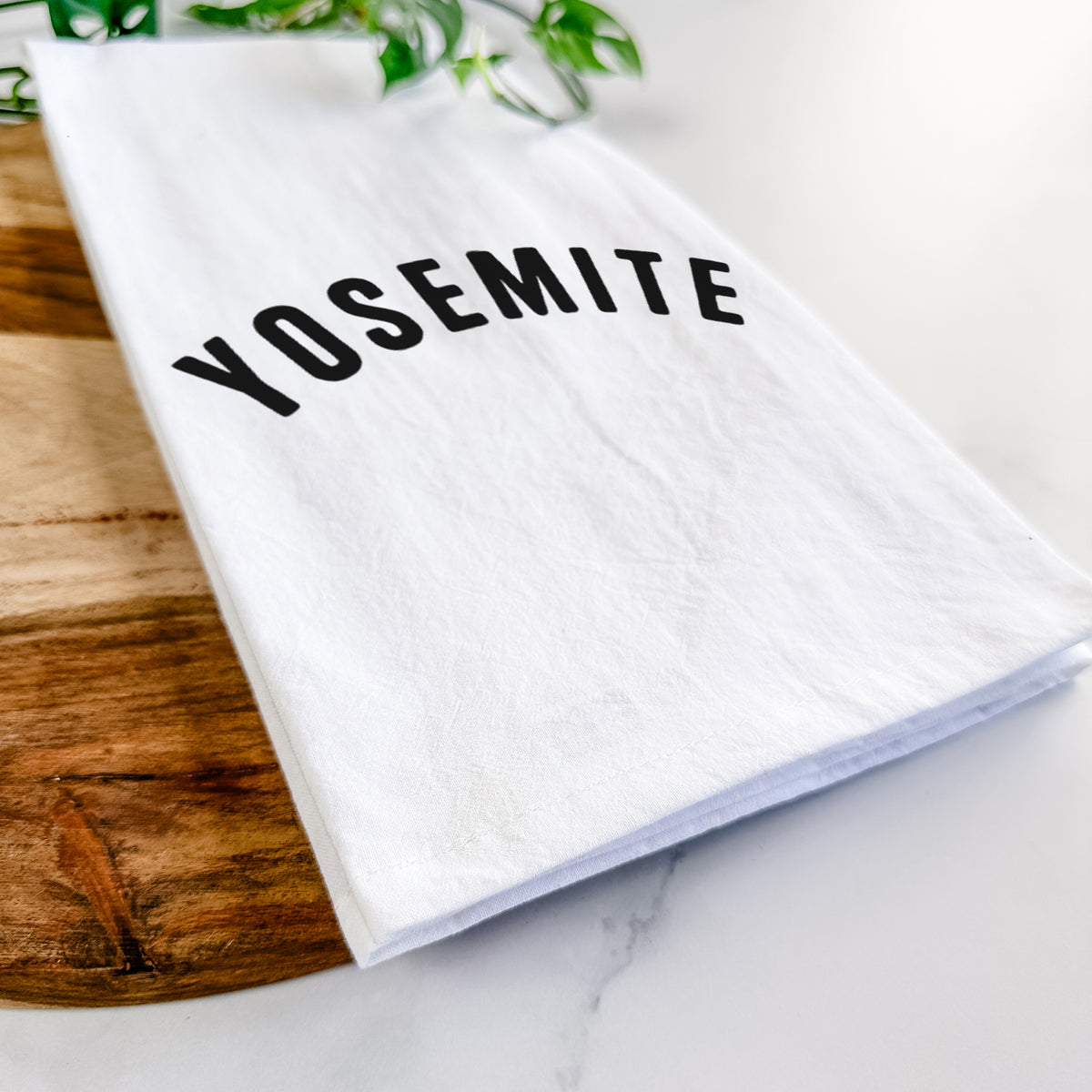 Close-up of a folded white tea towel on a wooden board, showing the word Yosemite in bold black letters arched across the chest.