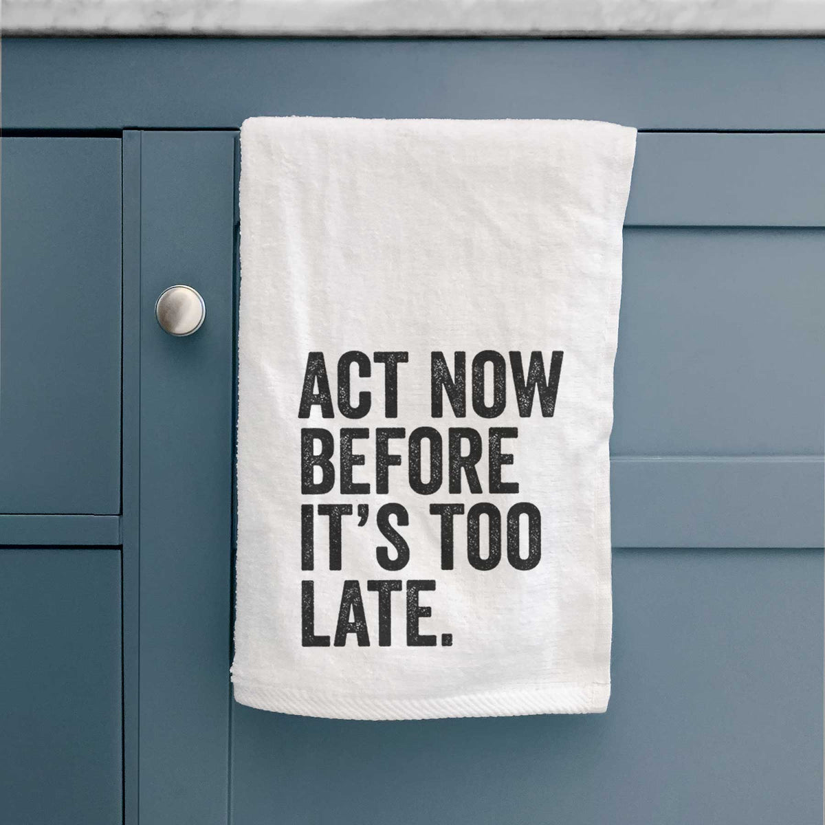 White hand towel printed with bold black words reading Act Now Before It's Too Late, hanging on a cabinet door.