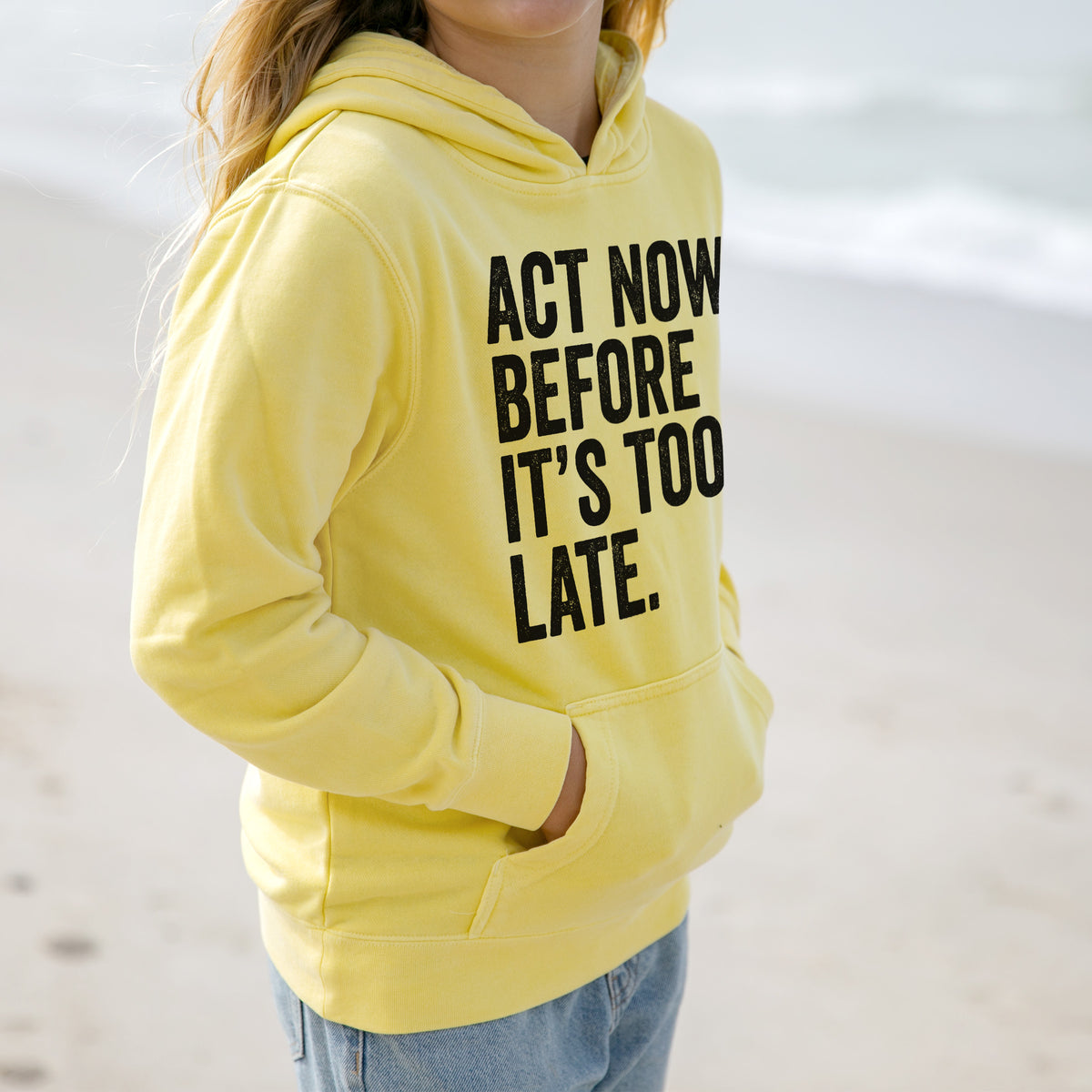 Girl wearing a youth pigment dyed hoodie printed with bold black words reading Act Now Before It's Too Late.