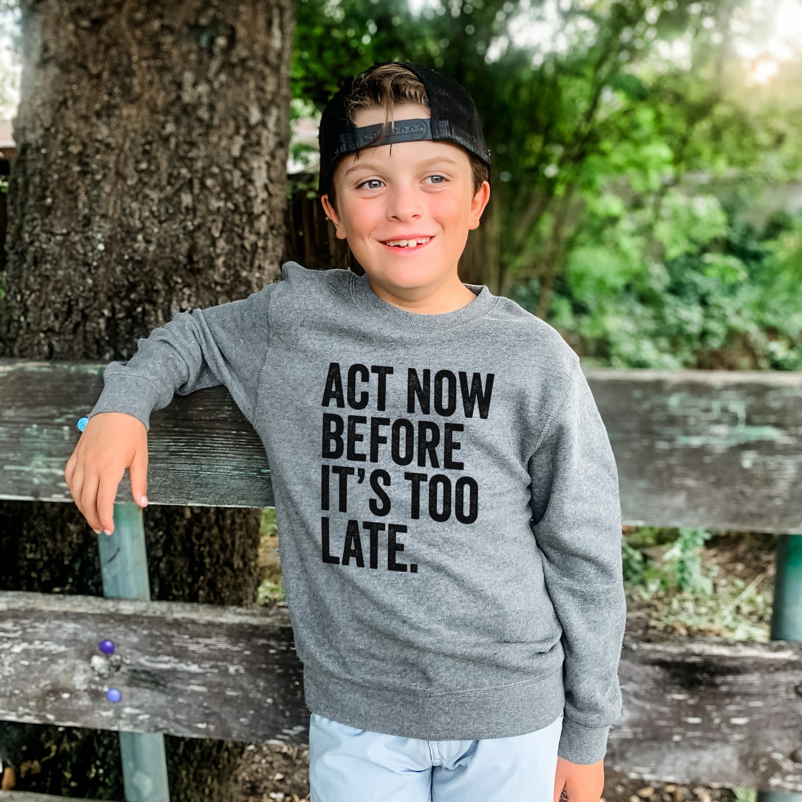 Boy wearing a youth crewneck sweatshirt printed with bold black words reading Act Now Before It's Too Late, outdoors.