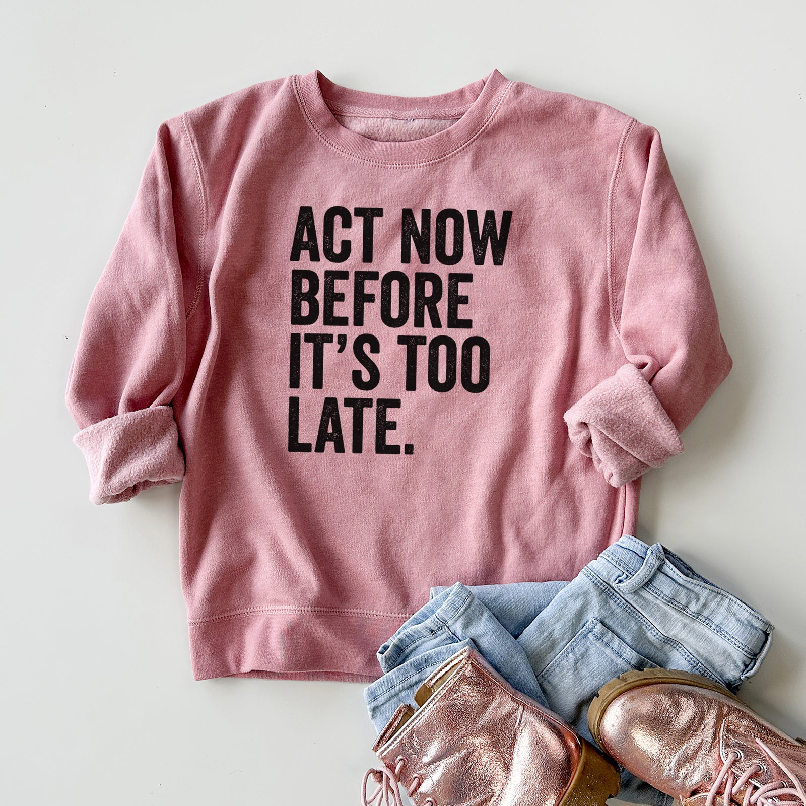 Boy wearing a youth crewneck sweatshirt printed with bold black words reading Act Now Before It's Too Late, outdoors.