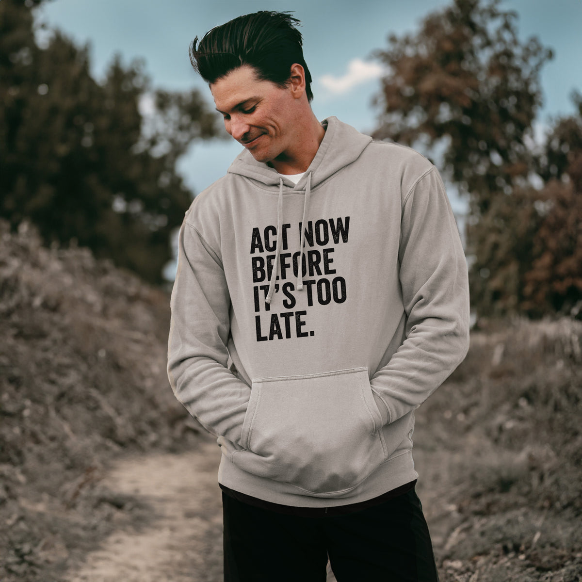 Man wearing a cement grey pigment dyed hoodie printed with bold black words reading Act Now Before It's Too Late.