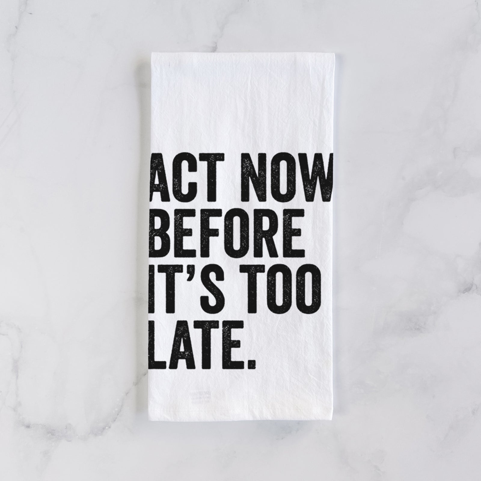 White cotton tea towel printed with bold black words reading Act Now Before It's Too Late, on a marble counter beside a round wooden cutting board and greenery.