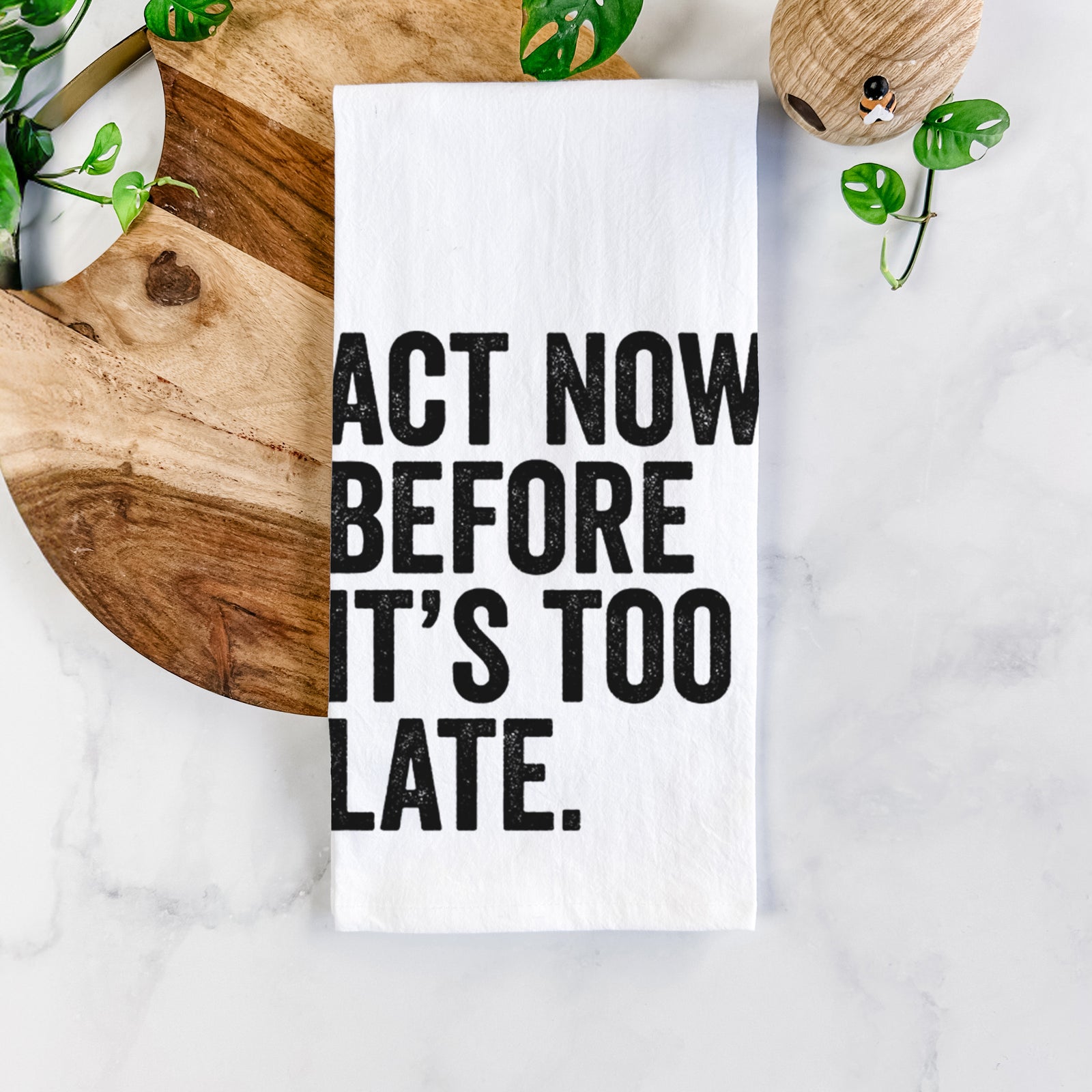 White cotton tea towel printed with bold black words reading Act Now Before It's Too Late, on a marble counter beside a round wooden cutting board and greenery.