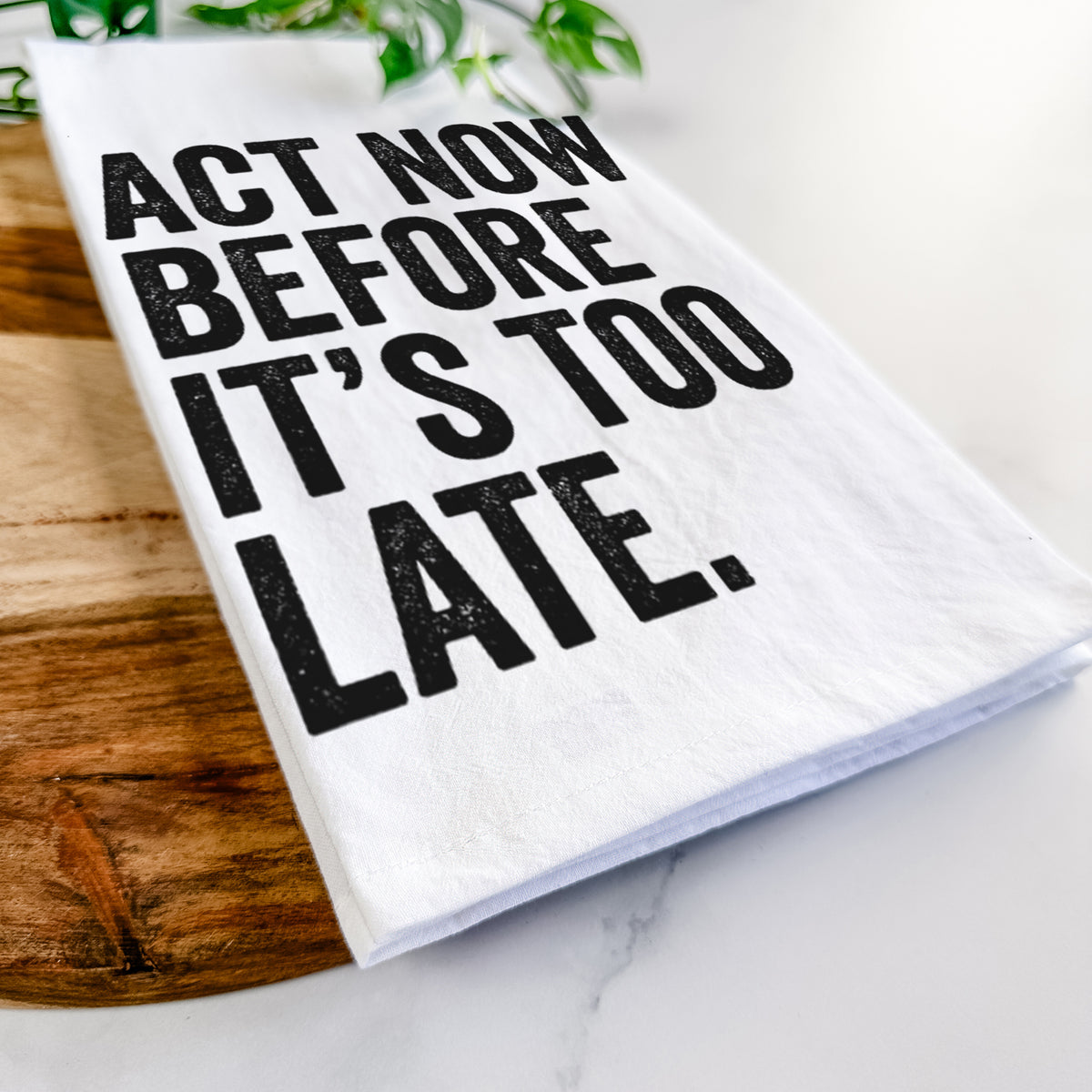 Close-up of a folded white tea towel on a wooden board, showing bold black words reading Act Now Before It's Too Late.