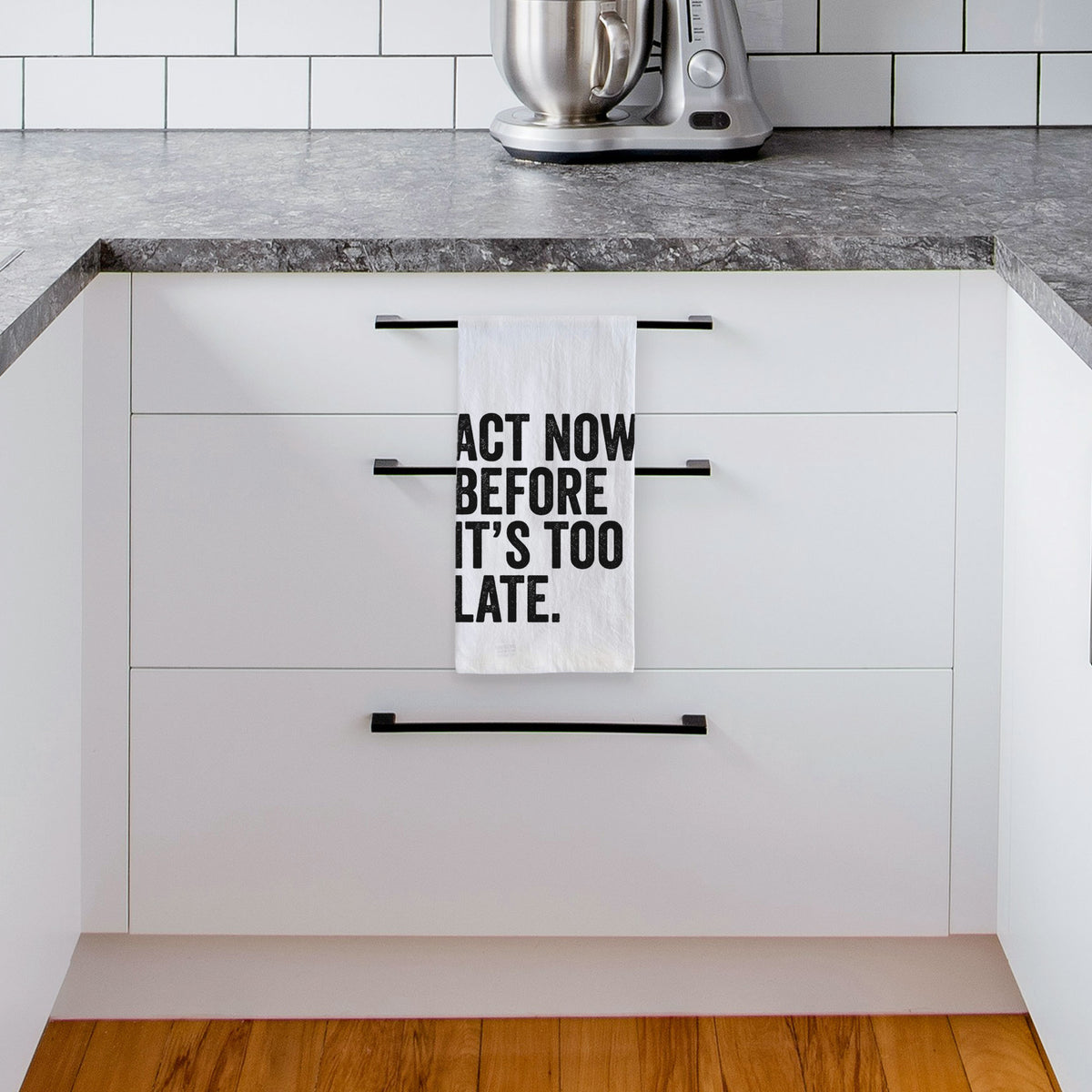 White tea towel printed with bold black words reading Act Now Before It's Too Late, hanging from a drawer handle in a bright white kitchen.