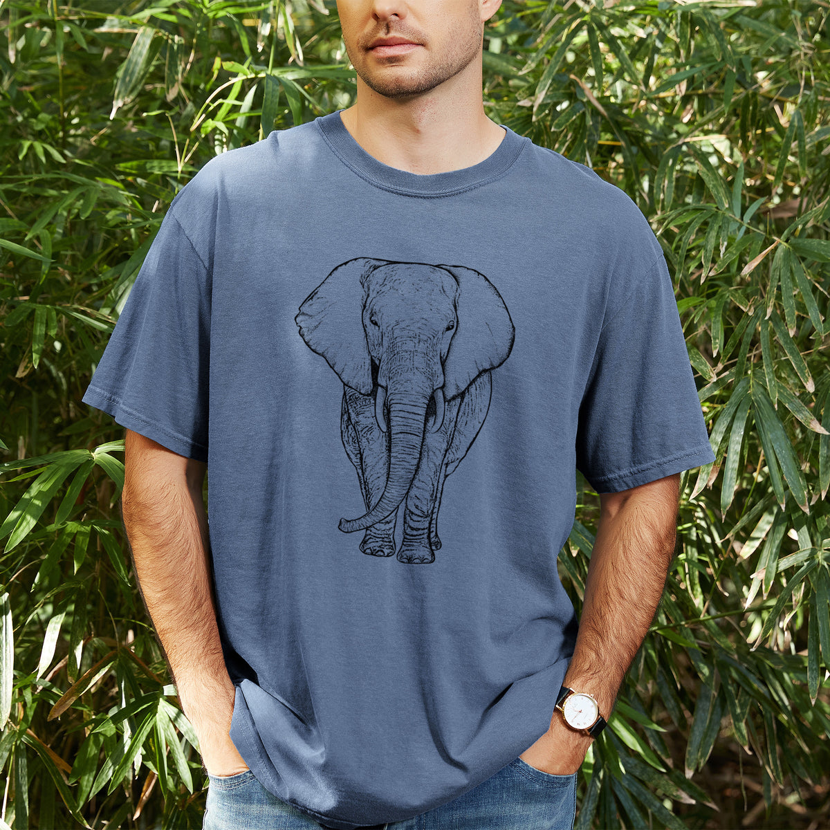 Man wearing a Comfort Colors t-shirt printed with a black ink line drawing of an African elephant facing forward with large ears and tusks, outdoors.