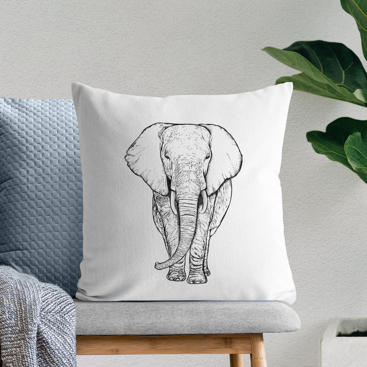 White pillow cover printed with a black ink line drawing of an African elephant facing forward with large ears and tusks, on a bench beside plants.