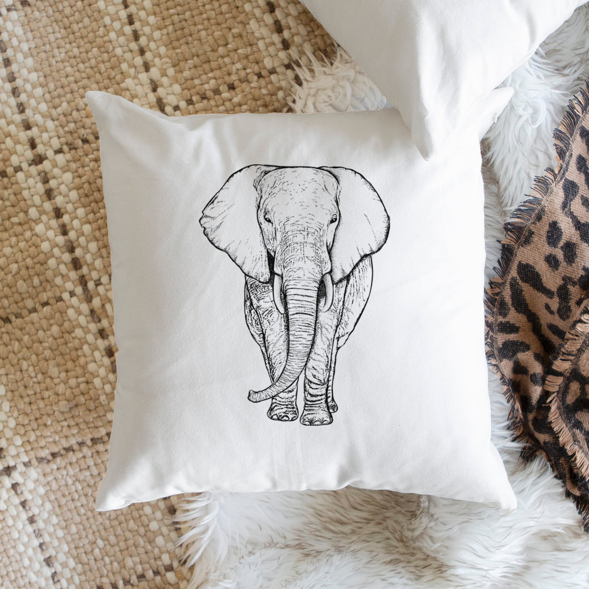 White pillow cover printed with a black ink line drawing of an African elephant facing forward with large ears and tusks, on a woven chair.
