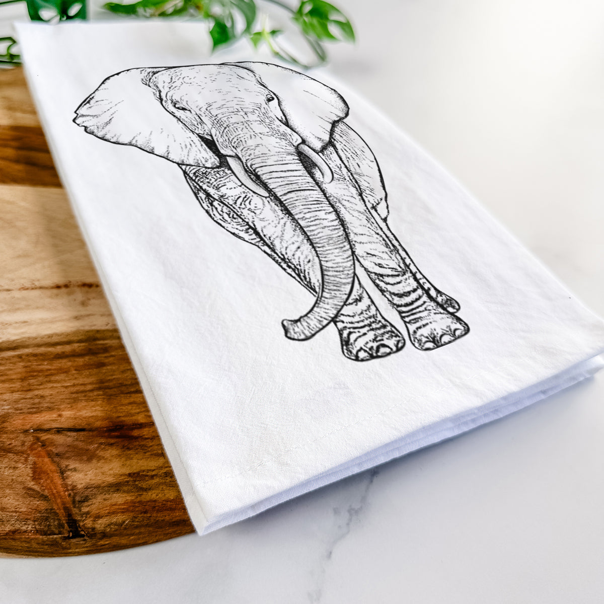 Close-up of a folded white tea towel on a wooden board, showing a black ink line drawing of an African elephant facing forward with large ears and tusks.