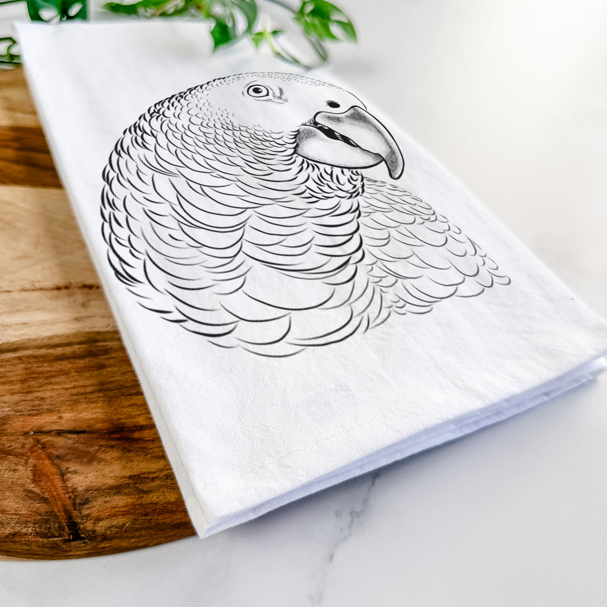 Close-up of a folded white tea towel on a wooden board, showing a detailed black ink portrait of an African grey parrot's head.