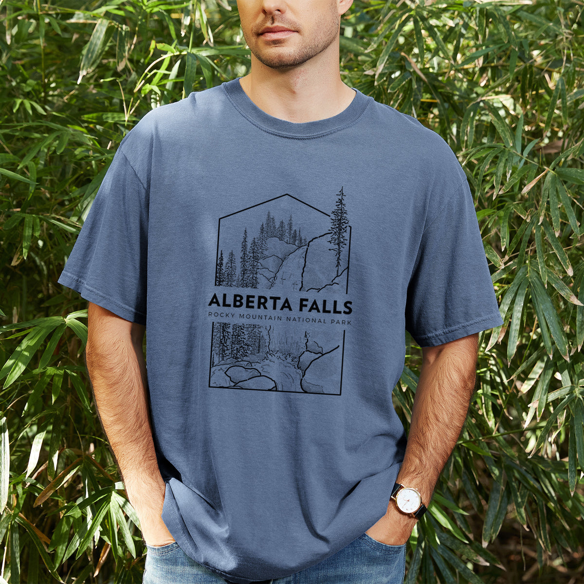Man wearing a Comfort Colors t-shirt printed with a black line art badge of Alberta Falls with pine trees, with the words Alberta Falls, Rocky Mountain National Park, outdoors.
