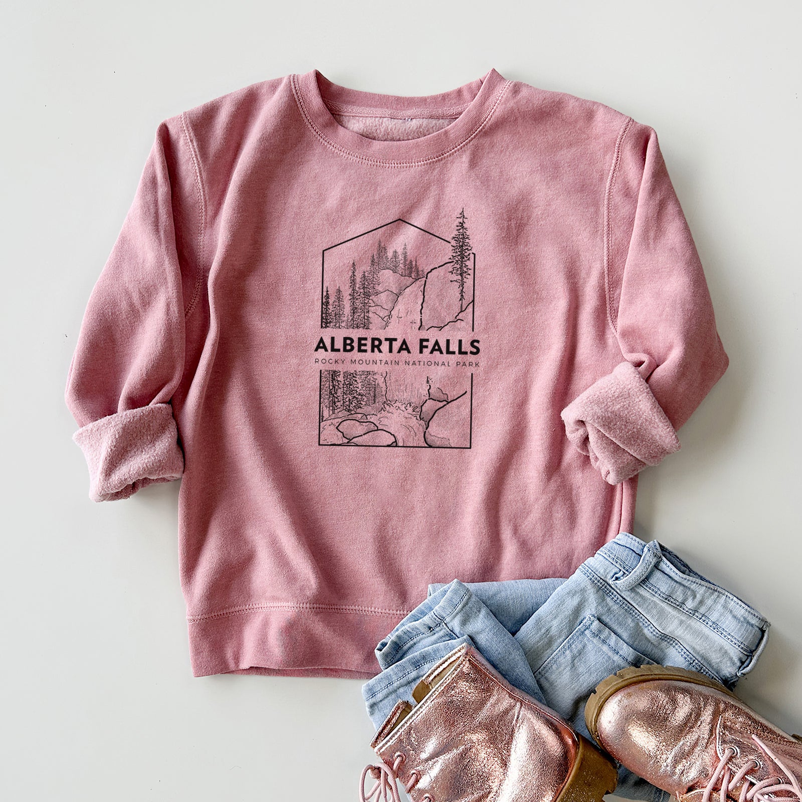 Boy wearing a youth crewneck sweatshirt printed with a black line art badge of Alberta Falls with pine trees, with the words Alberta Falls, Rocky Mountain National Park, outdoors.