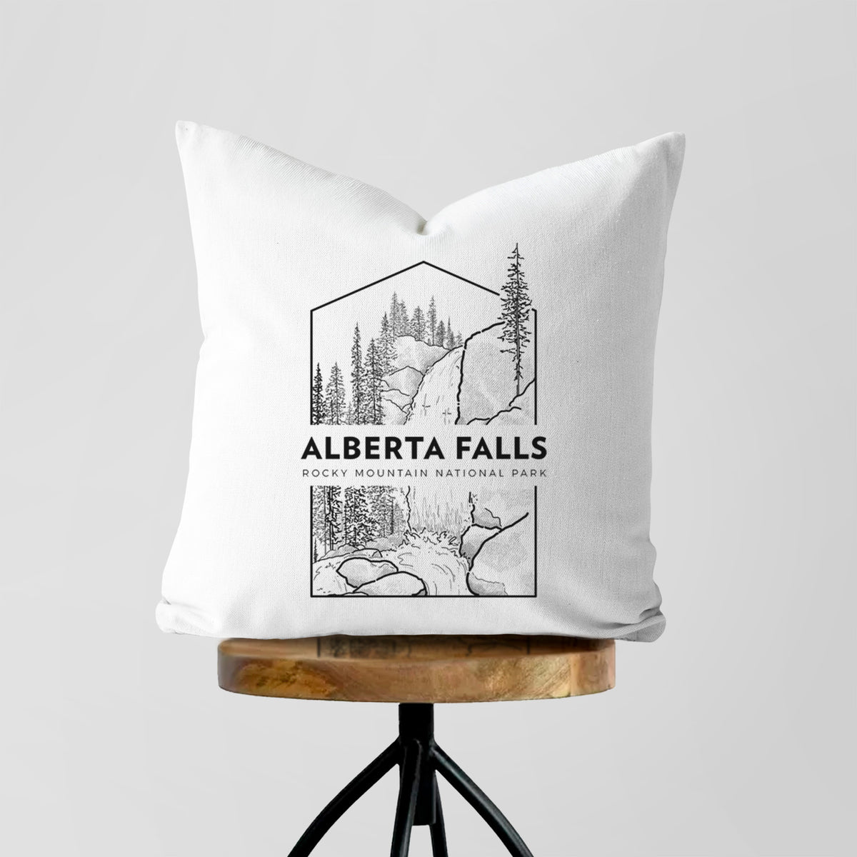 White pillow cover printed with a black line art badge of Alberta Falls with pine trees, with the words Alberta Falls, Rocky Mountain National Park, on a wooden stool.