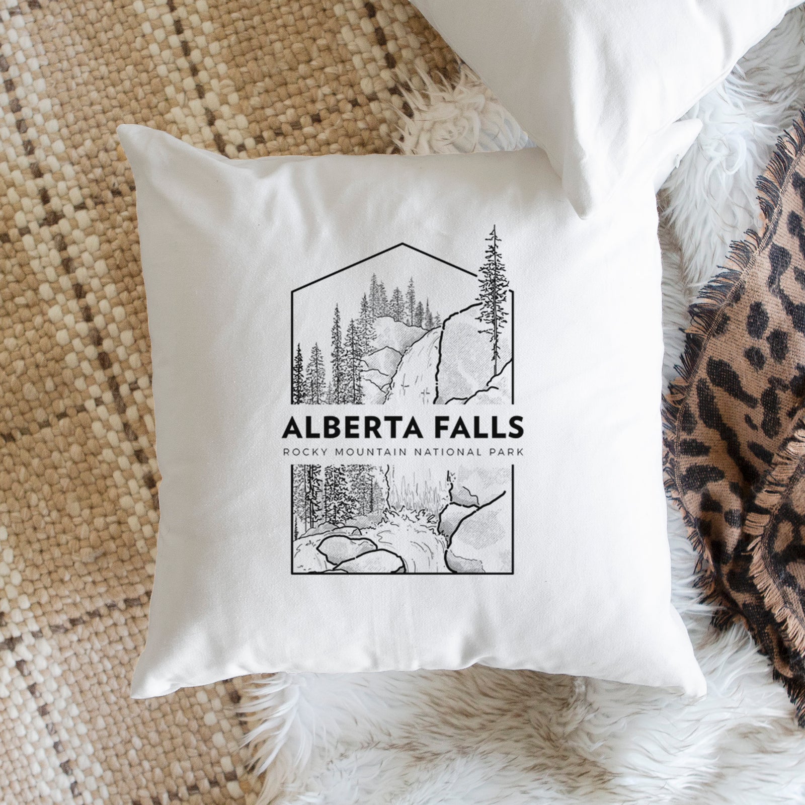 White pillow cover printed with a black line art badge of Alberta Falls with pine trees, with the words Alberta Falls, Rocky Mountain National Park, on a bench beside plants.