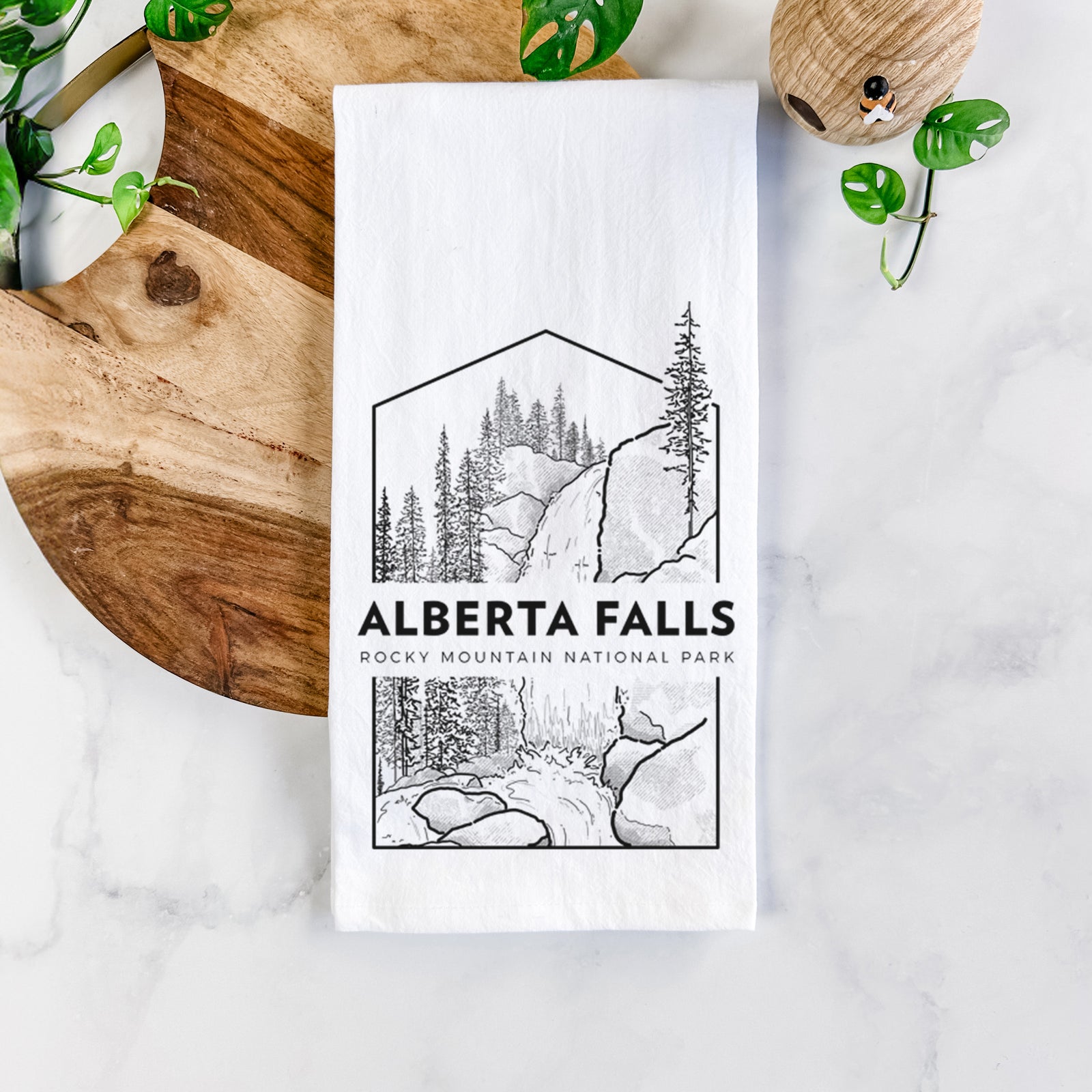 White cotton tea towel printed with a black line art badge of Alberta Falls with pine trees, with the words Alberta Falls, Rocky Mountain National Park, on a marble counter beside a round wooden cutting board and greenery.