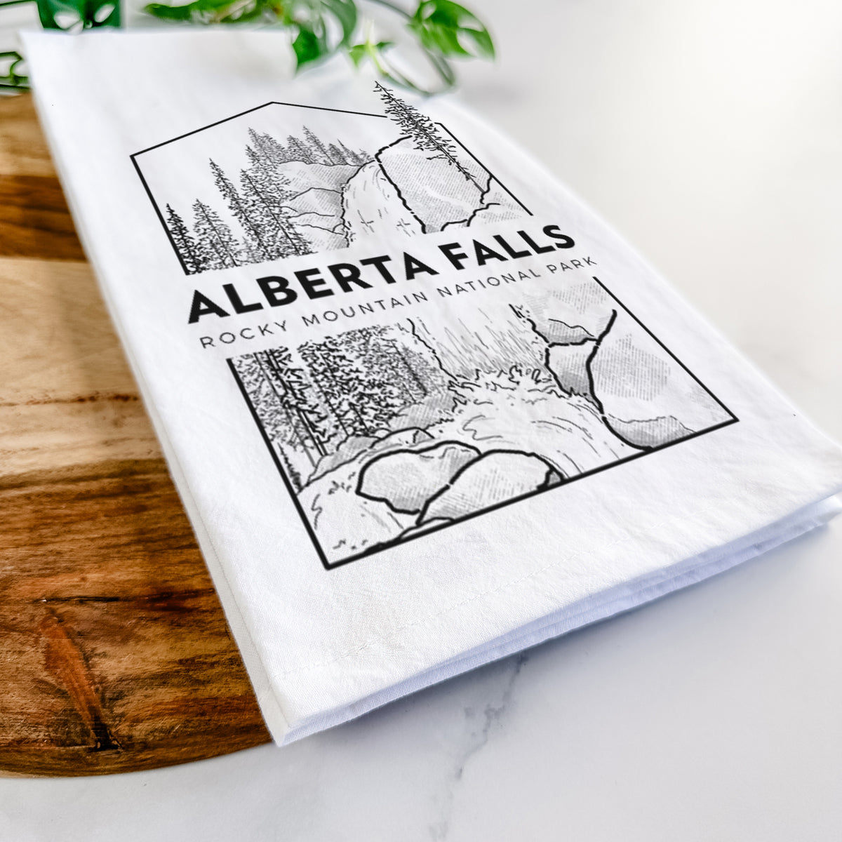 Close-up of a folded white tea towel on a wooden board, showing a black line art badge of Alberta Falls with pine trees, with the words Alberta Falls, Rocky Mountain National Park.