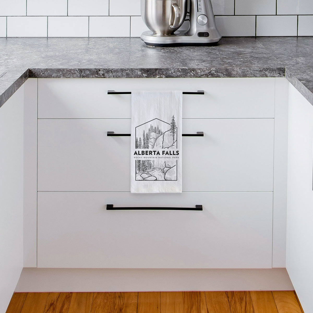 White tea towel printed with a black line art badge of Alberta Falls with pine trees, with the words Alberta Falls, Rocky Mountain National Park, hanging from a drawer handle in a bright white kitchen.