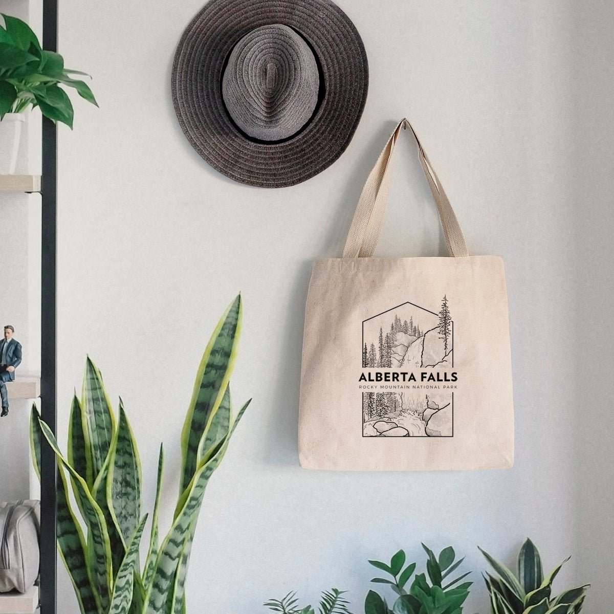 Canvas tote bag printed with a black line art badge of Alberta Falls with pine trees, with the words Alberta Falls, Rocky Mountain National Park, hanging on a wall hook below a hat, next to potted plants.