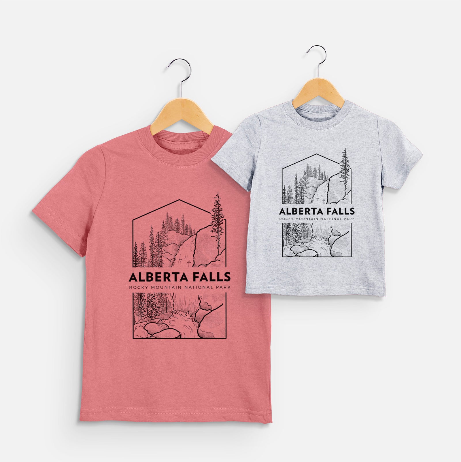 Two youth t-shirts in different colors on hangers, printed with a black line art badge of Alberta Falls with pine trees, with the words Alberta Falls, Rocky Mountain National Park.