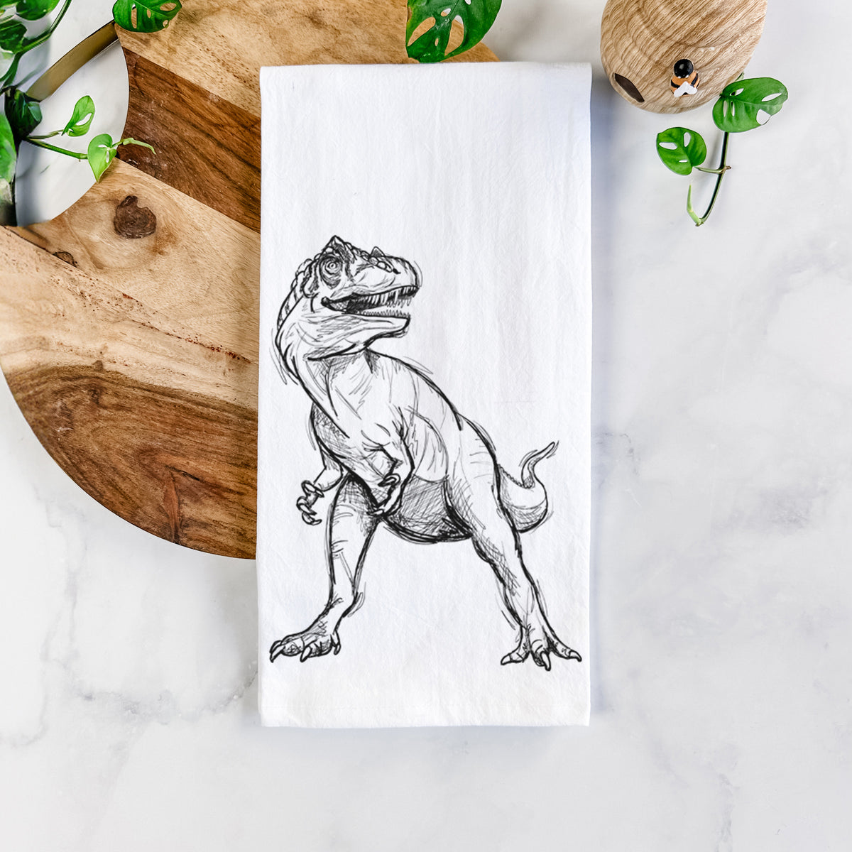 White cotton tea towel printed with a black ink illustration of an Allosaurus walking, on a marble counter beside a round wooden cutting board and greenery.
