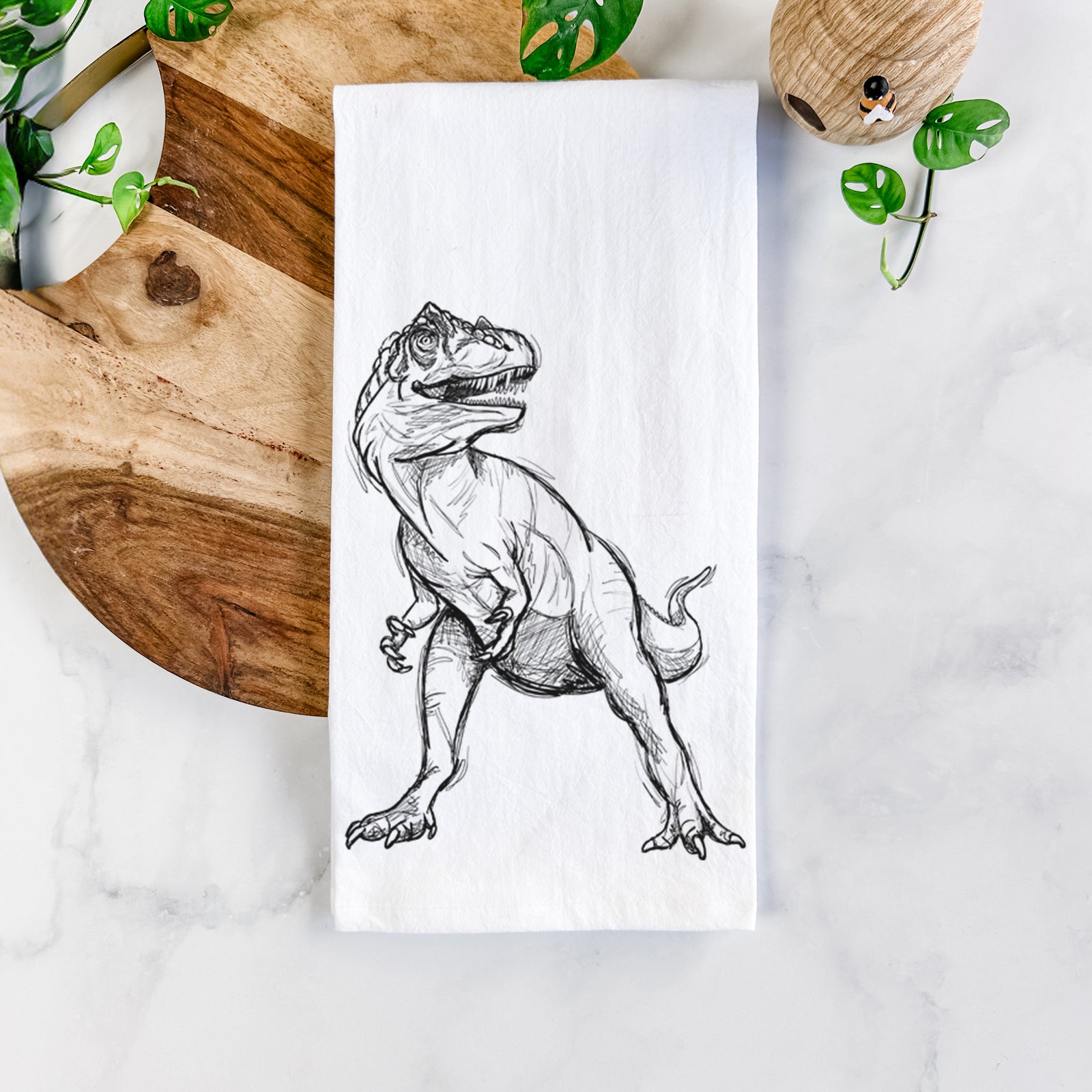 White cotton tea towel printed with a black ink illustration of an Allosaurus walking, on a marble counter beside a round wooden cutting board and greenery.