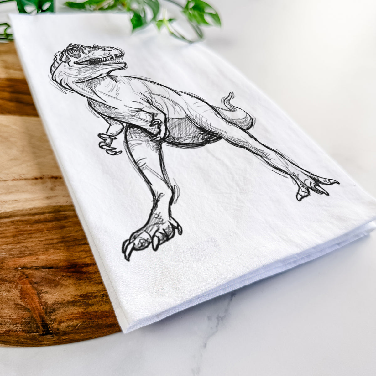 Close-up of a folded white tea towel on a wooden board, showing a black ink illustration of an Allosaurus walking.