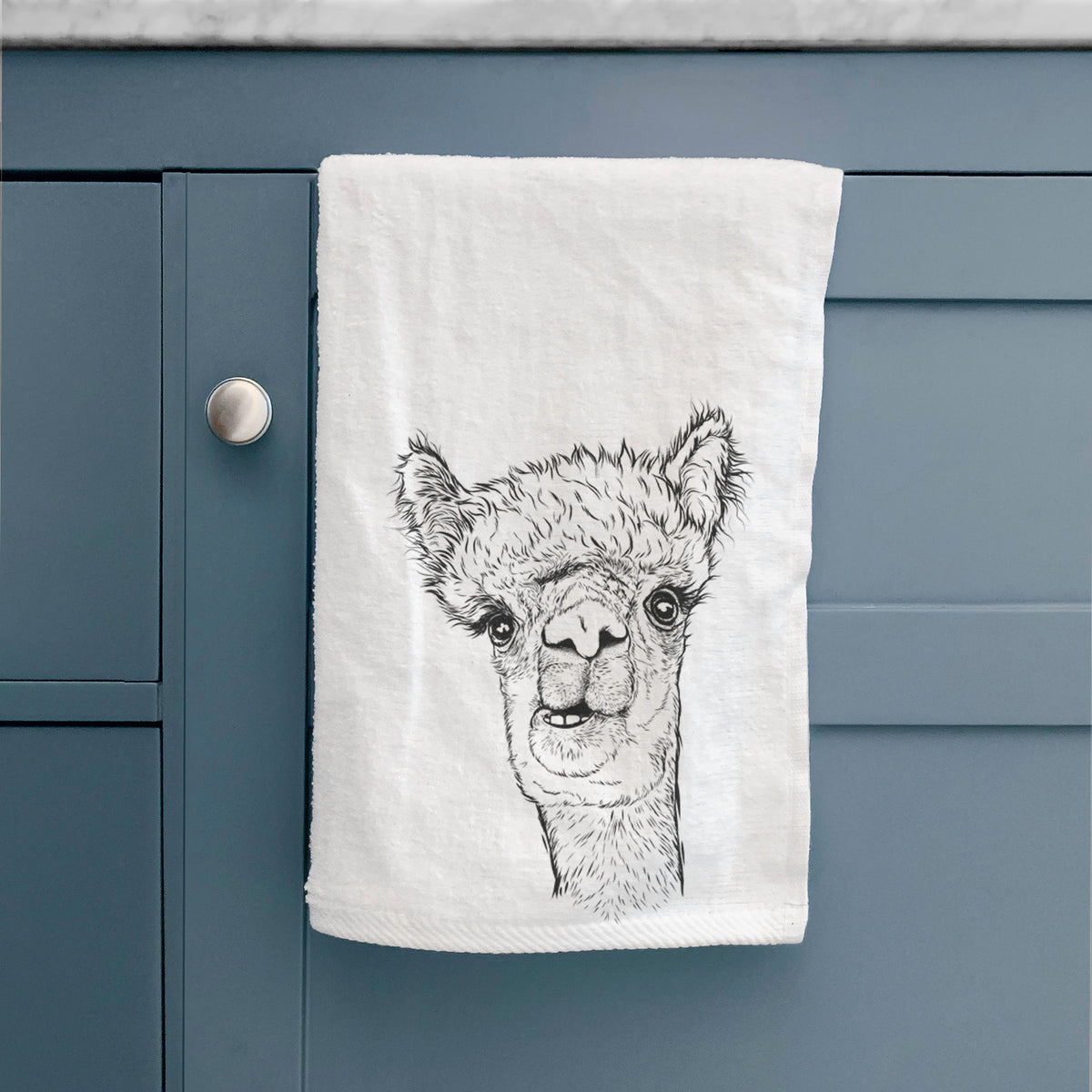 White hand towel printed with a detailed black ink portrait of a fluffy alpaca's head and neck, hanging on a cabinet door.