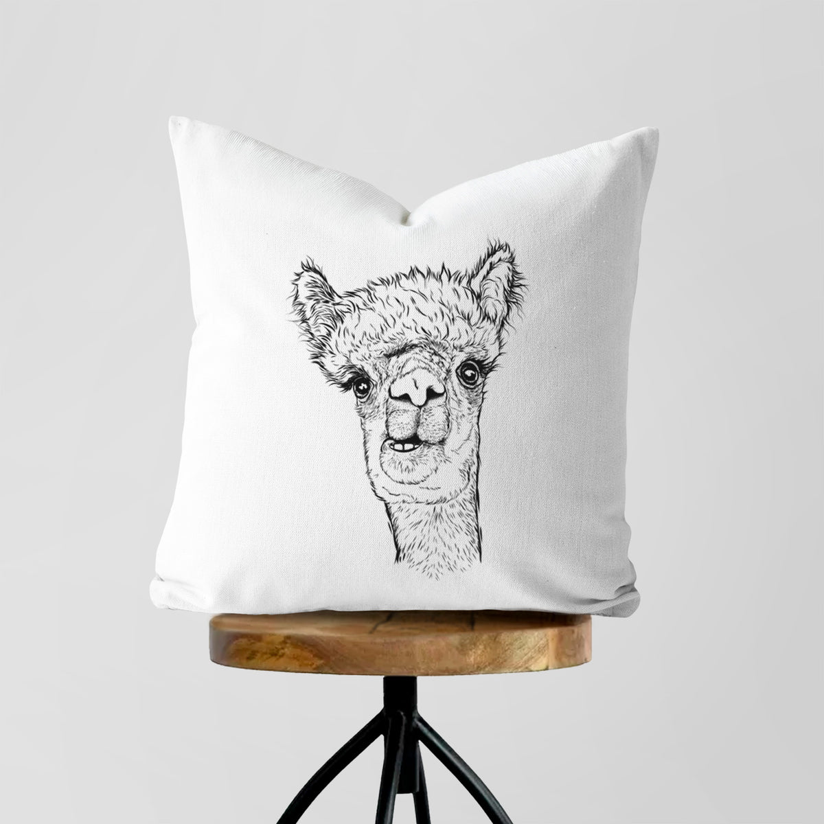White pillow cover printed with a detailed black ink portrait of a fluffy alpaca's head and neck, on a wooden stool.