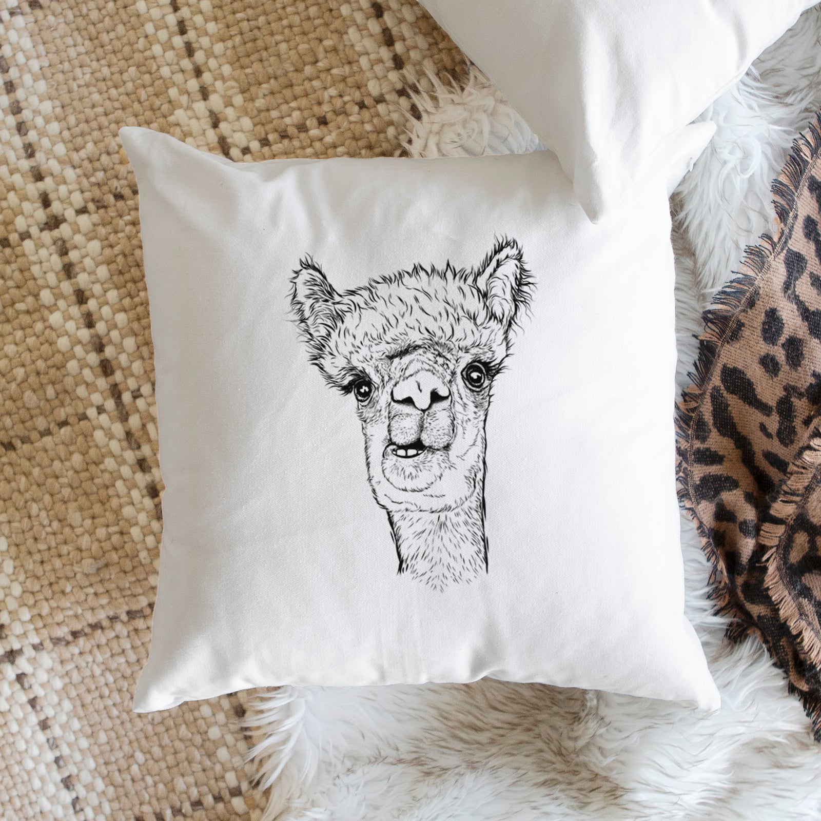 White pillow cover printed with a detailed black ink portrait of a fluffy alpaca's head and neck, on a bench beside plants.