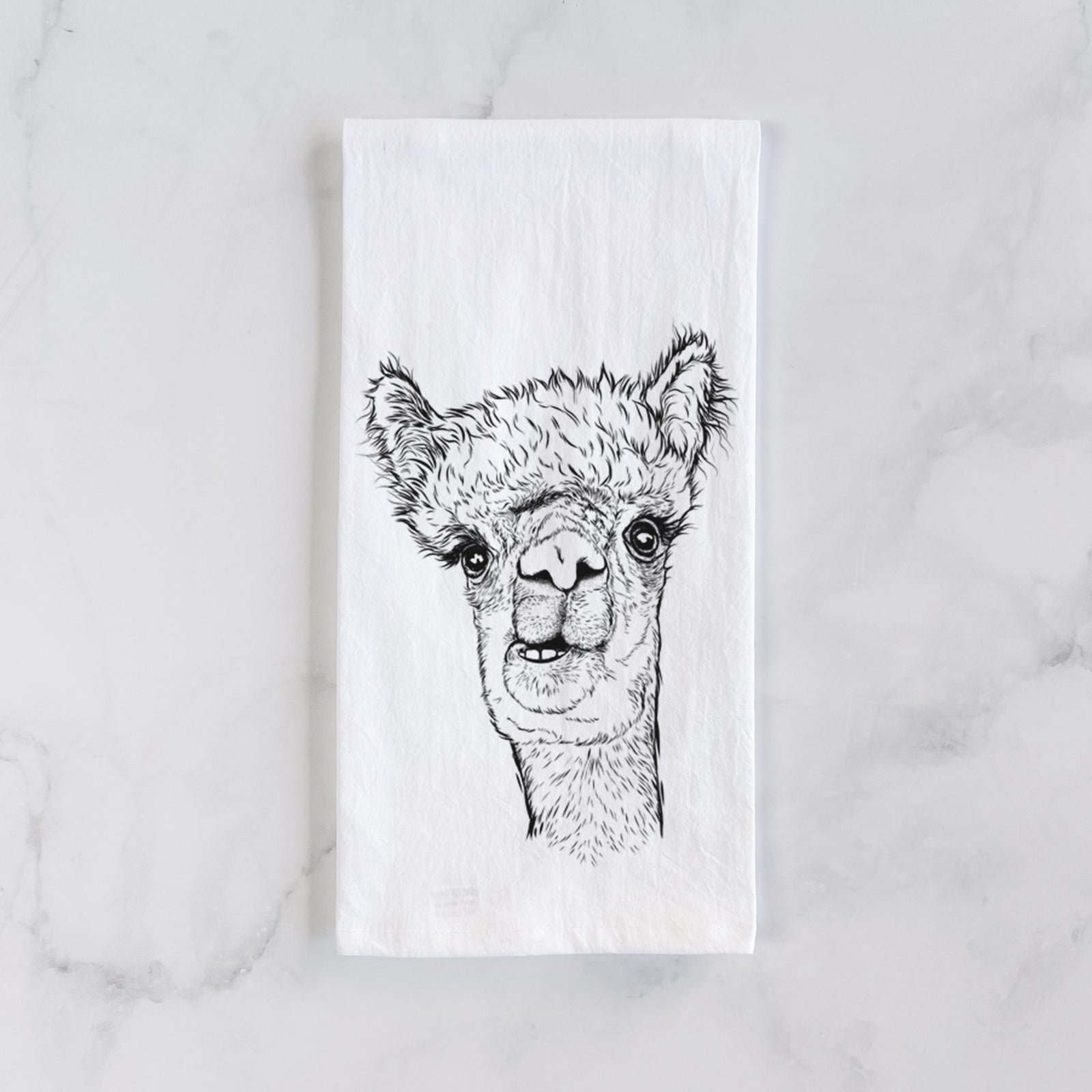 White cotton tea towel printed with a detailed black ink portrait of a fluffy alpaca's head and neck, on a marble counter beside a round wooden cutting board and greenery.