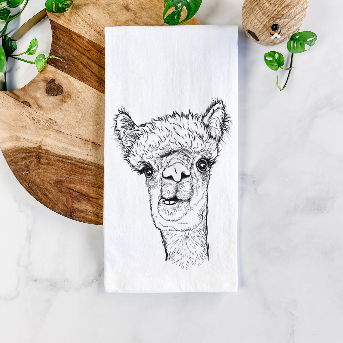 White cotton tea towel printed with a detailed black ink portrait of a fluffy alpaca's head and neck, on a marble counter beside a round wooden cutting board and greenery.
