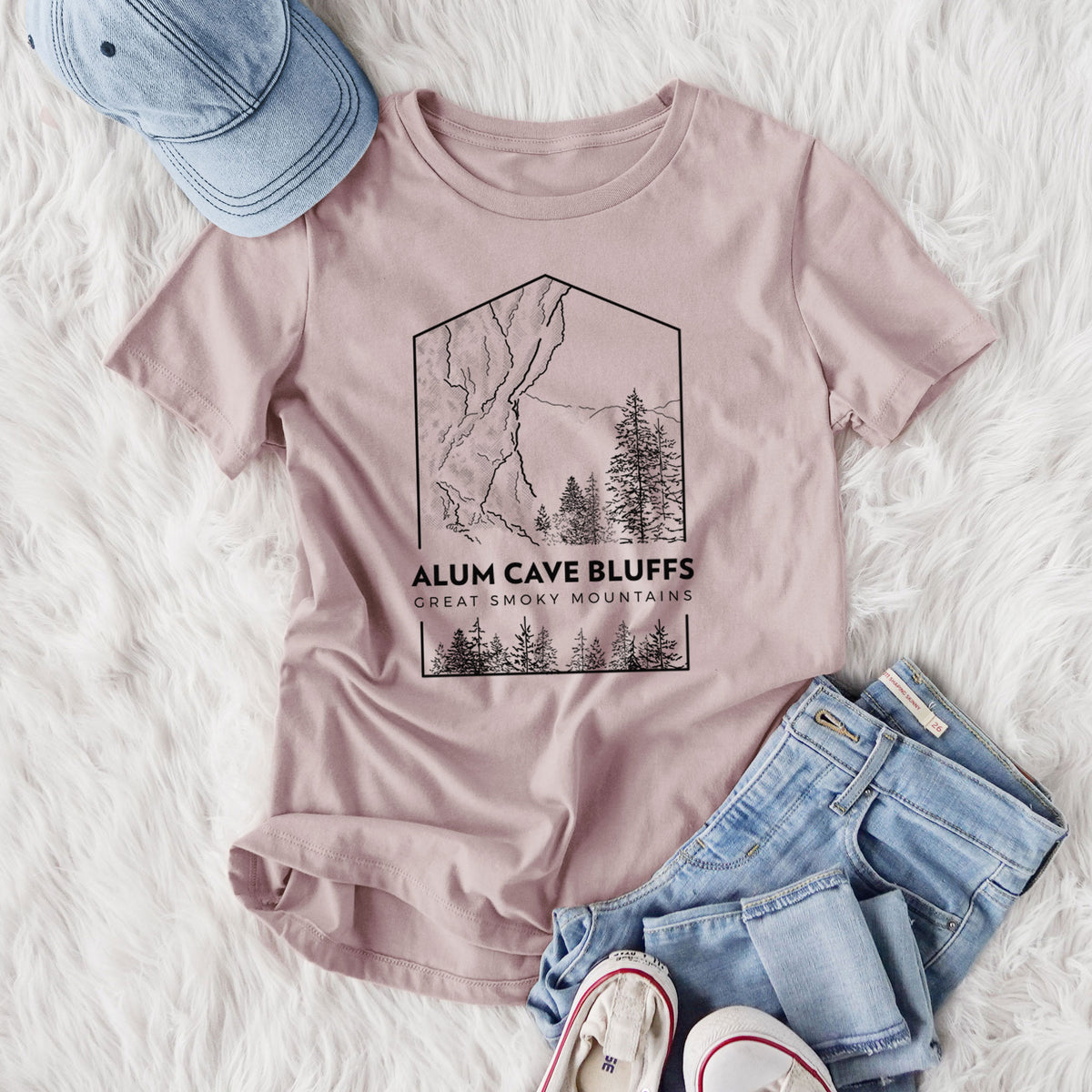 Women's relaxed fit cotton t-shirt printed with a black line art badge of Alum Cave Bluffs with pine trees, with the words Alum Cave Bluffs, Great Smoky Mountains, styled flat on a white faux fur rug with jeans.