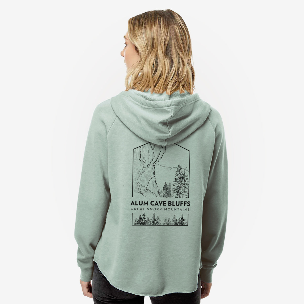Woman wearing a sage green women's Cali Wave zip-up sweatshirt printed with a black line art badge of Alum Cave Bluffs with pine trees, with the words Alum Cave Bluffs, Great Smoky Mountains, seen from behind.