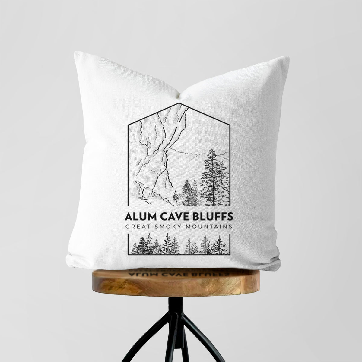 White pillow cover printed with a black line art badge of Alum Cave Bluffs with pine trees, with the words Alum Cave Bluffs, Great Smoky Mountains, on a wooden stool.