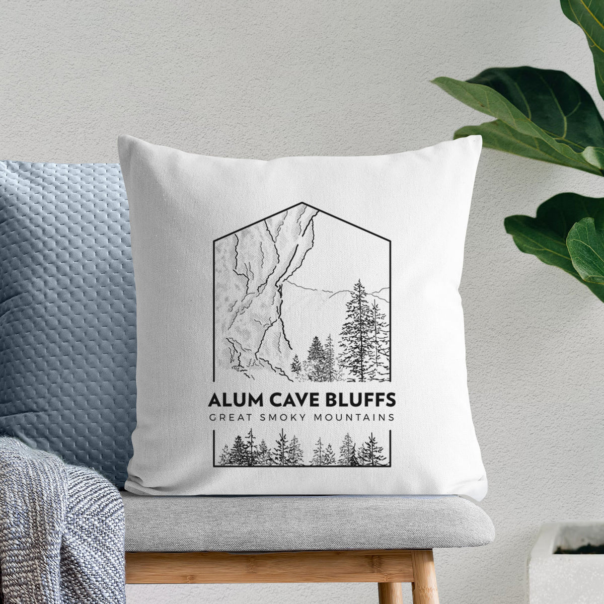White pillow cover printed with a black line art badge of Alum Cave Bluffs with pine trees, with the words Alum Cave Bluffs, Great Smoky Mountains, on a bench beside plants.