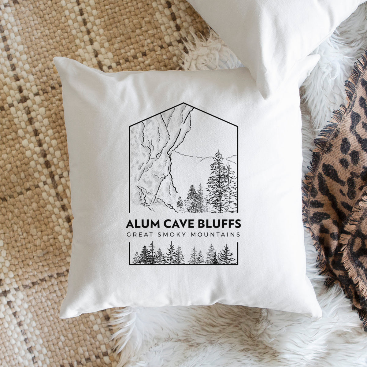 White pillow cover printed with a black line art badge of Alum Cave Bluffs with pine trees, with the words Alum Cave Bluffs, Great Smoky Mountains, on a woven chair.