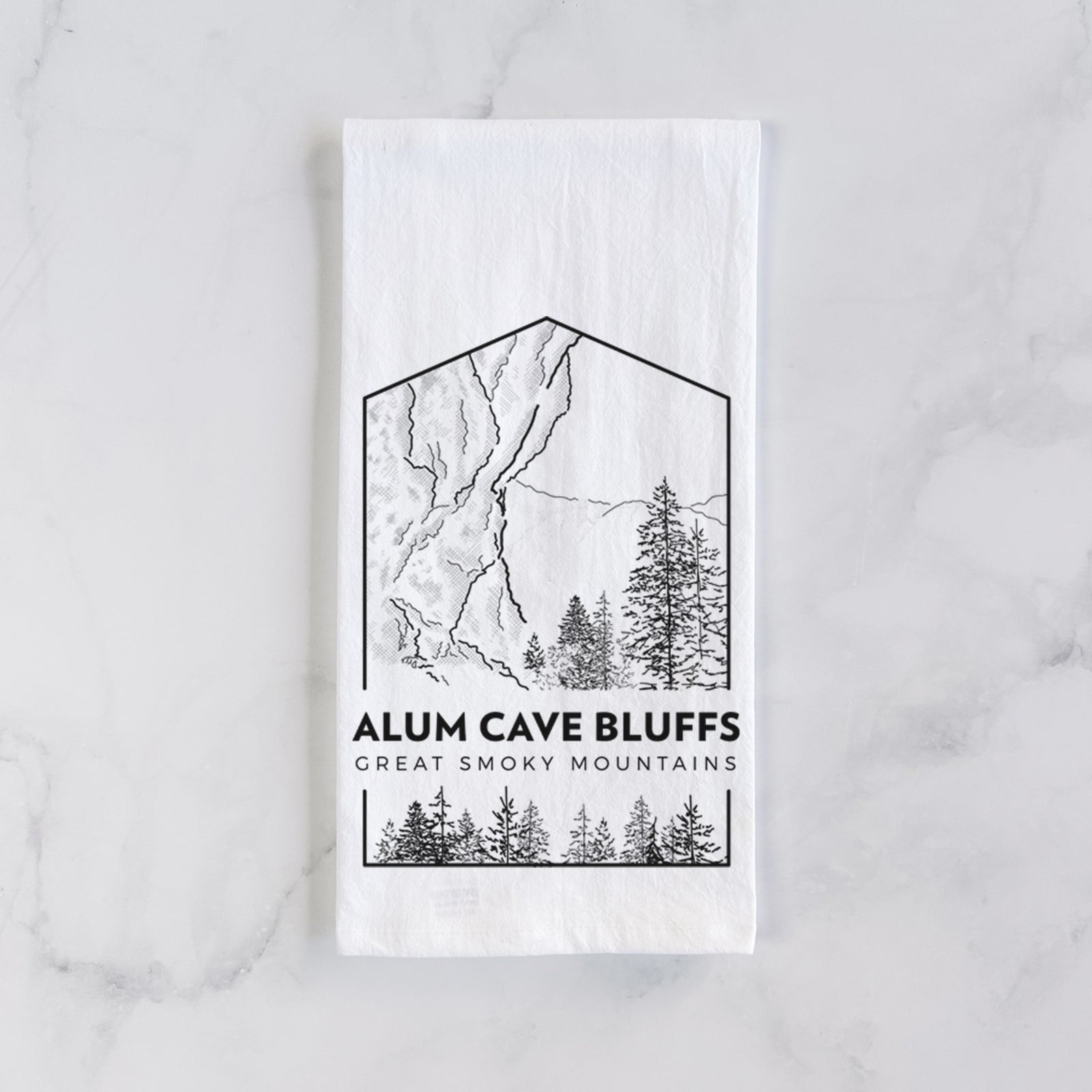 White cotton tea towel printed with a black line art badge of Alum Cave Bluffs with pine trees, with the words Alum Cave Bluffs, Great Smoky Mountains, on a marble counter beside a round wooden cutting board and greenery.
