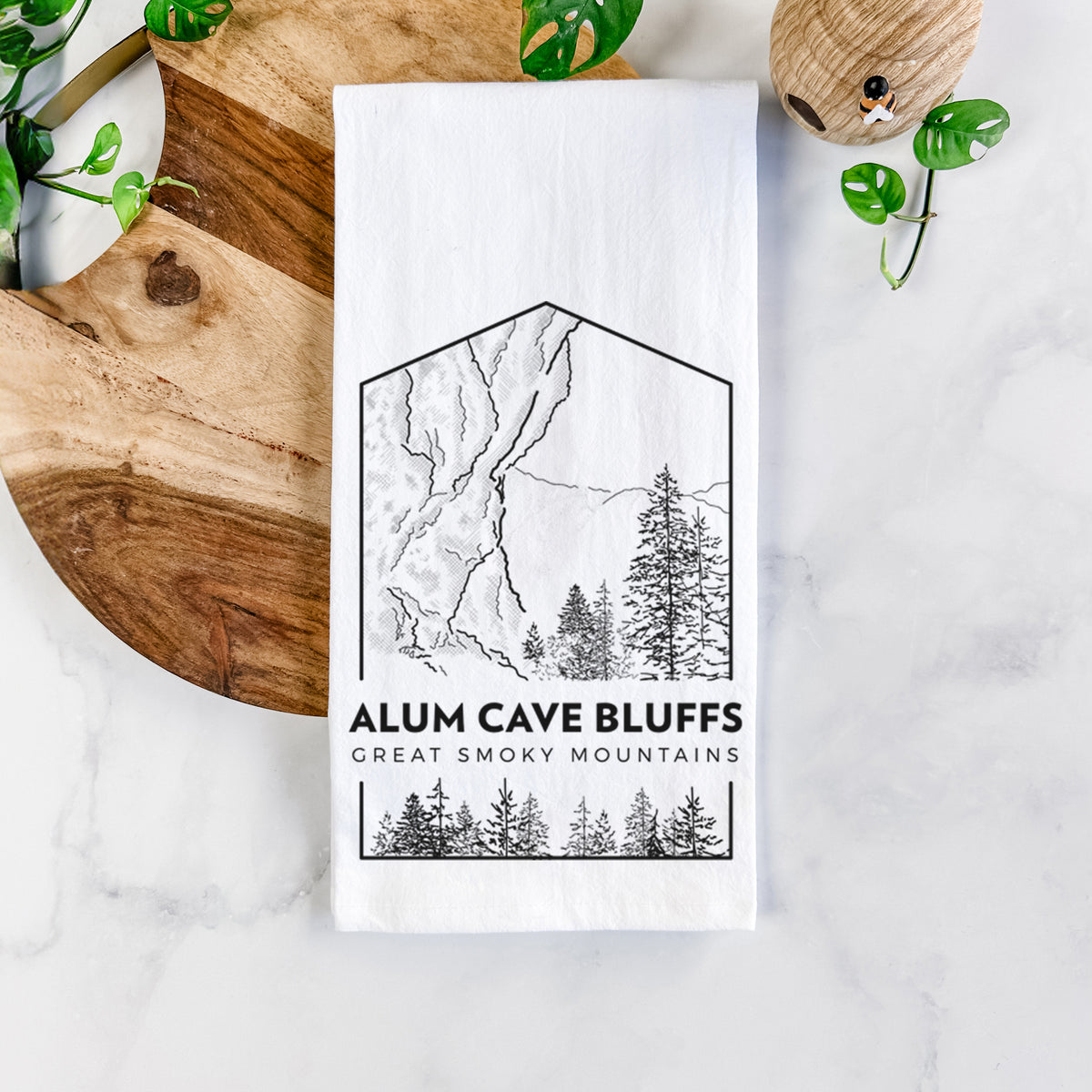 White cotton tea towel printed with a black line art badge of Alum Cave Bluffs with pine trees, with the words Alum Cave Bluffs, Great Smoky Mountains, on a marble counter beside a round wooden cutting board and greenery.