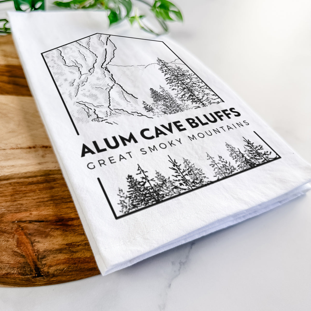 Close-up of a folded white tea towel on a wooden board, showing a black line art badge of Alum Cave Bluffs with pine trees, with the words Alum Cave Bluffs, Great Smoky Mountains.