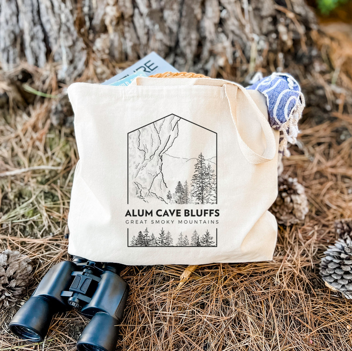 Canvas tote bag printed with a black line art badge of Alum Cave Bluffs with pine trees, with the words Alum Cave Bluffs, Great Smoky Mountains, resting on dry grass beside a pair of binoculars.
