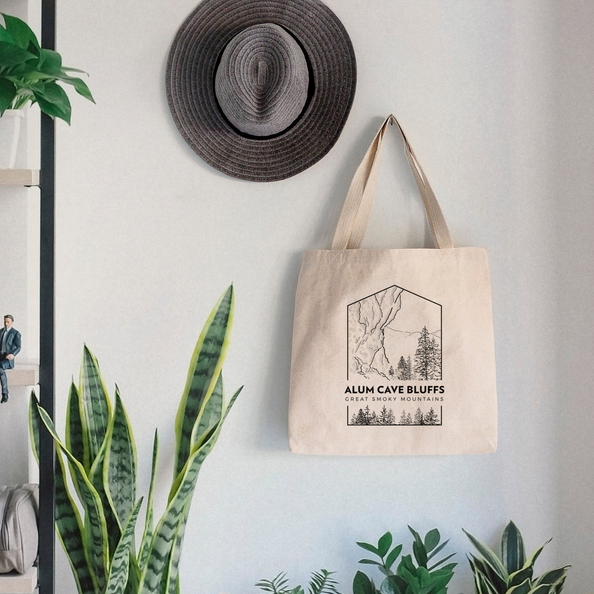 Canvas tote bag printed with a black line art badge of Alum Cave Bluffs with pine trees, with the words Alum Cave Bluffs, Great Smoky Mountains, hanging on a wall hook below a hat, next to potted plants.